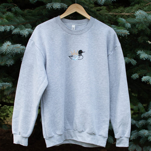 A light heather gray crewneck sweatshirt hanging on a wooden hanger against a background of green pine trees. The sweatshirt features a small embroidered loon design on the center chest.
