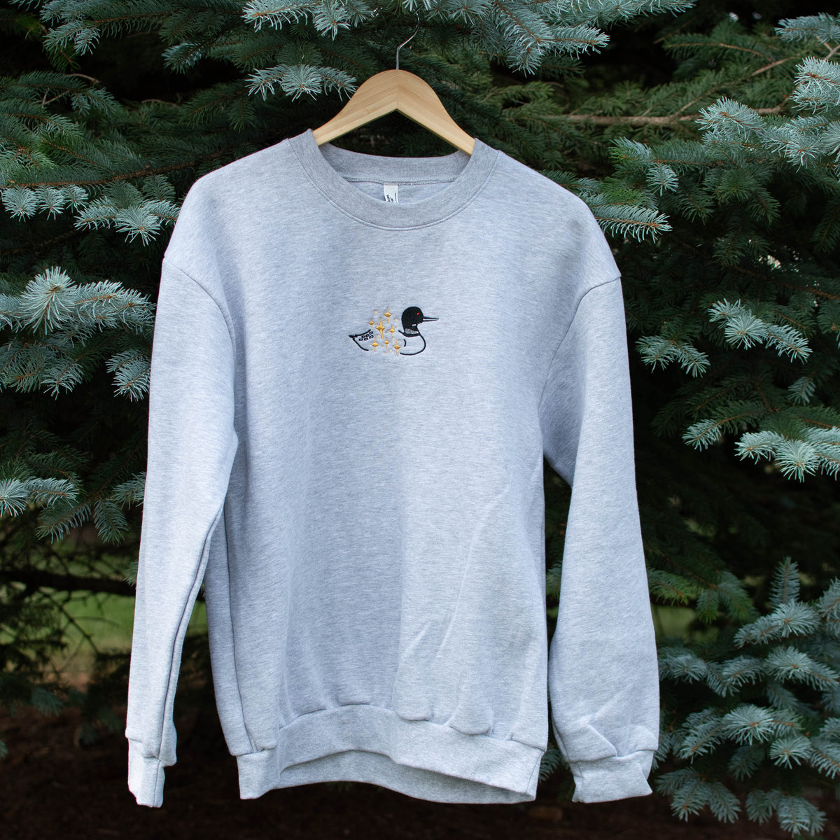 A light heather gray crewneck sweatshirt hanging on a wooden hanger against a background of green pine trees. The sweatshirt features a small embroidered loon design on the center chest.
