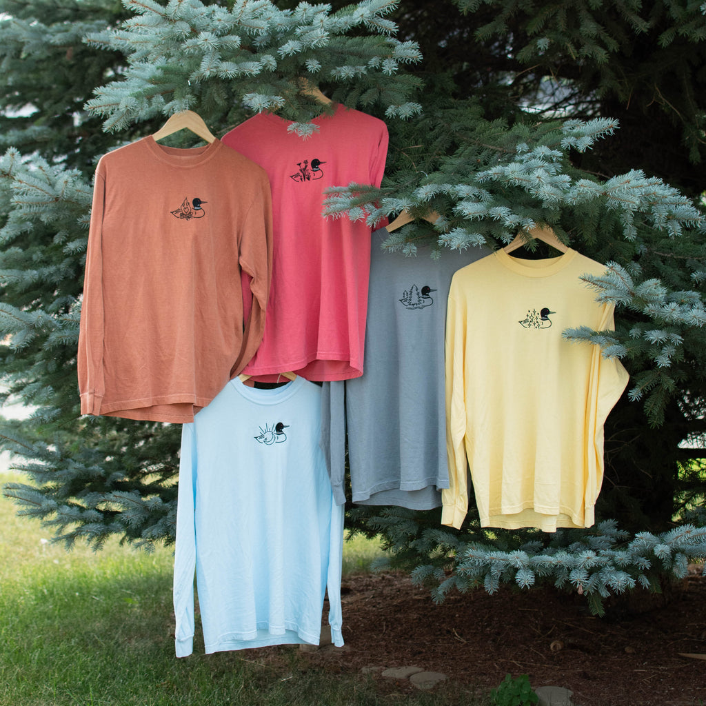 Five long-sleeve crewneck shirts in different colors (from top left to right and bottom: yam, watermelon, granite, butter, and chambray) are hanging on wooden hangers from the branches of a pine tree outdoors.