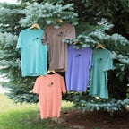 Five short sleeve t-shirts in different colors (from top left to right and bottom: seafoam, espresso, violet, light green, and terracotta) are hanging on wooden hangers from the branches of a pine tree outdoors.