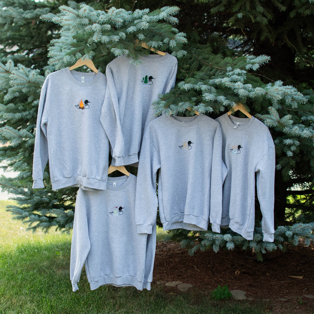 Five light gray crewneck sweatshirts with small embroidered graphics, hanging on wooden hangers from the branches of a green evergreen tree outdoors.