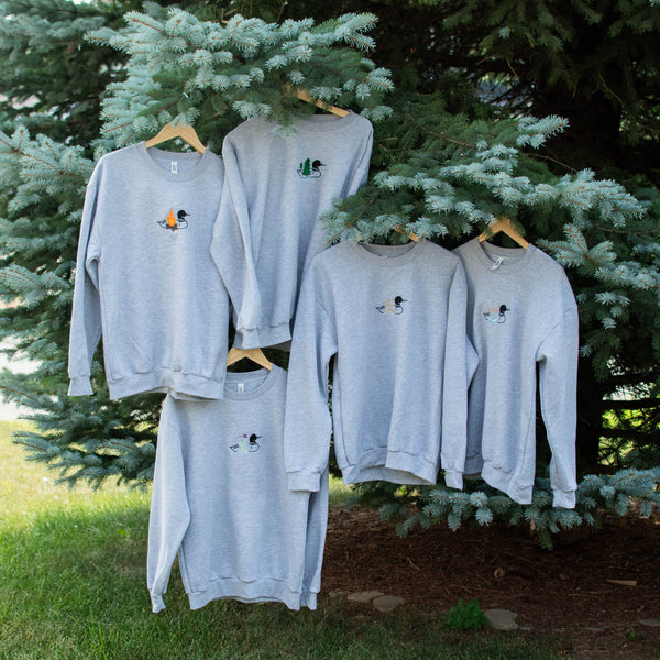 Five light gray crewneck sweatshirts with small embroidered graphics, hanging on wooden hangers from the branches of a green evergreen tree outdoors.