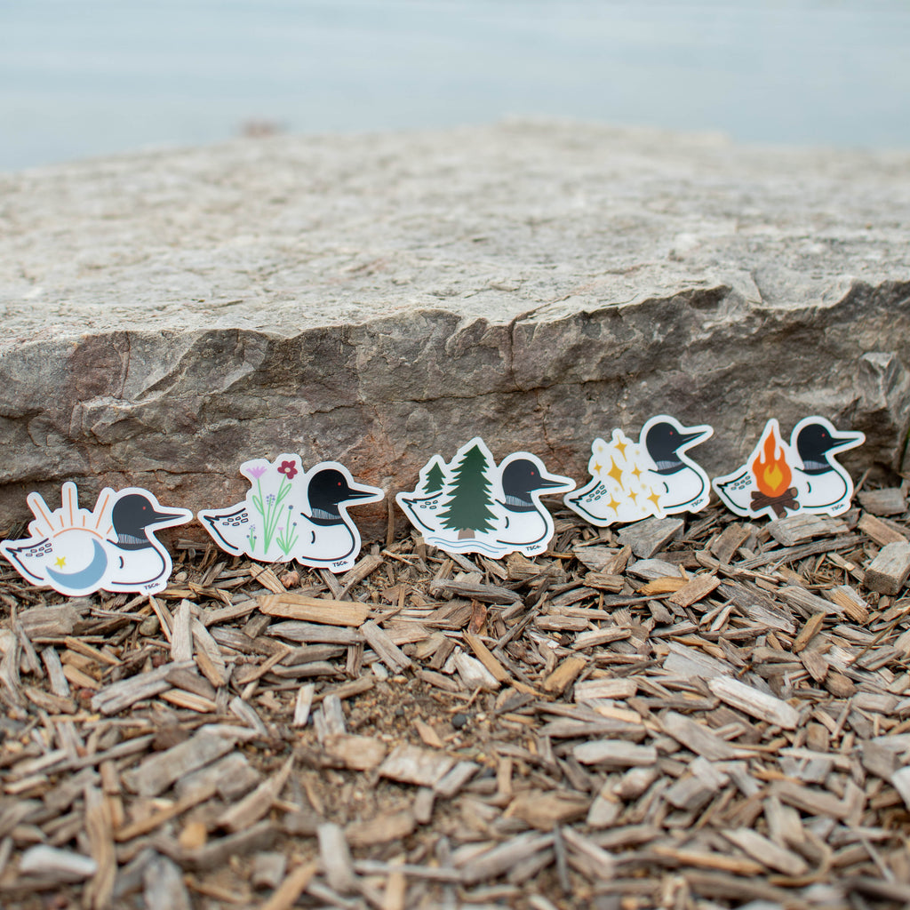 Six loon-shaped stickers with different nature-themed designs are lined up on a bed of wood mulch in front of a large stone, with a blurred body of water in the background.