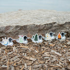 Six loon-shaped stickers with different nature-themed designs are lined up on a bed of wood mulch in front of a large stone, with a blurred body of water in the background.