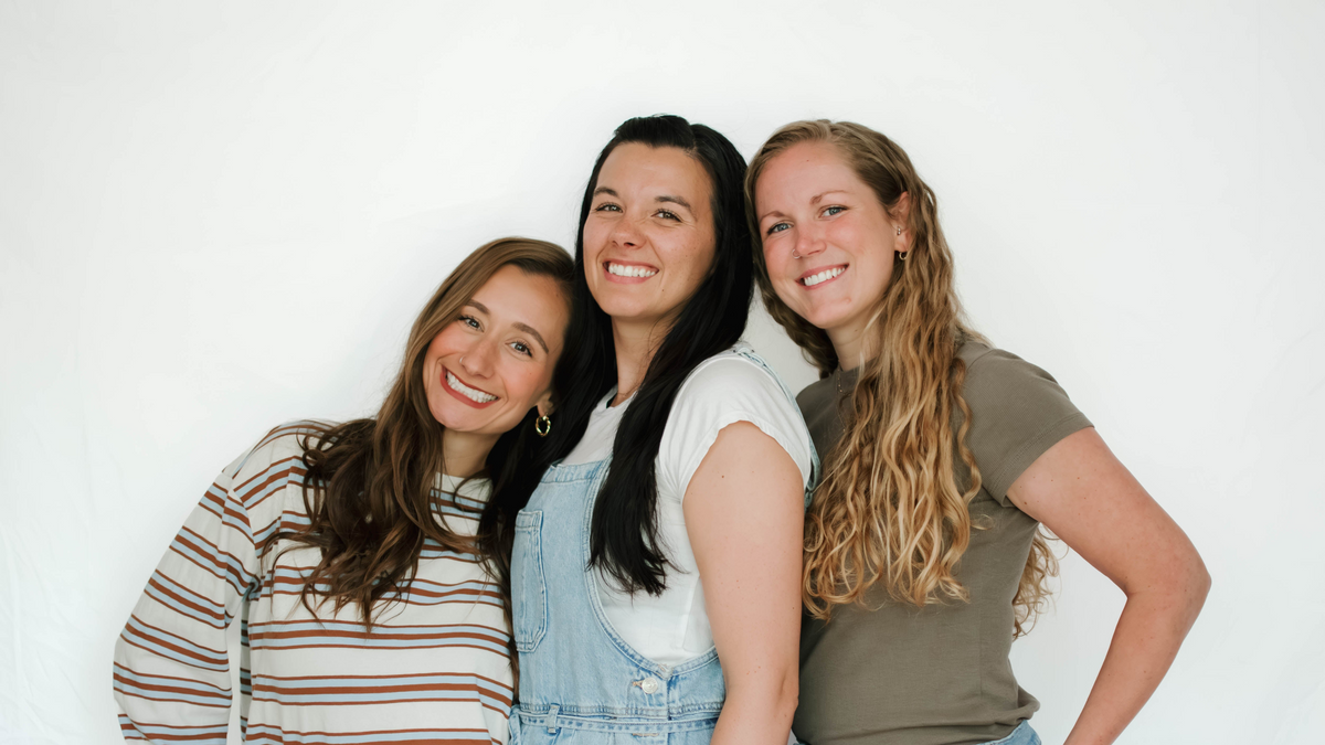 Three young women smiling and posing together against a plain white studio backdrop. The woman on the left wears a brown and white striped long-sleeve shirt, the woman in the center wears light blue denim overalls over a white top, and the woman on the right wears a muted brown short-sleeve t-shirt.