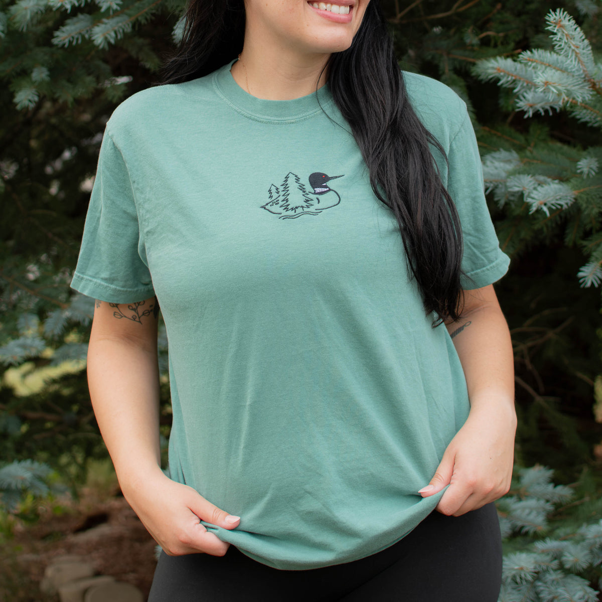 A woman wearing a light green colored crewneck sweatshirt with a small black embroidered loon design on the center chest, standing outdoors in front of green pine trees.