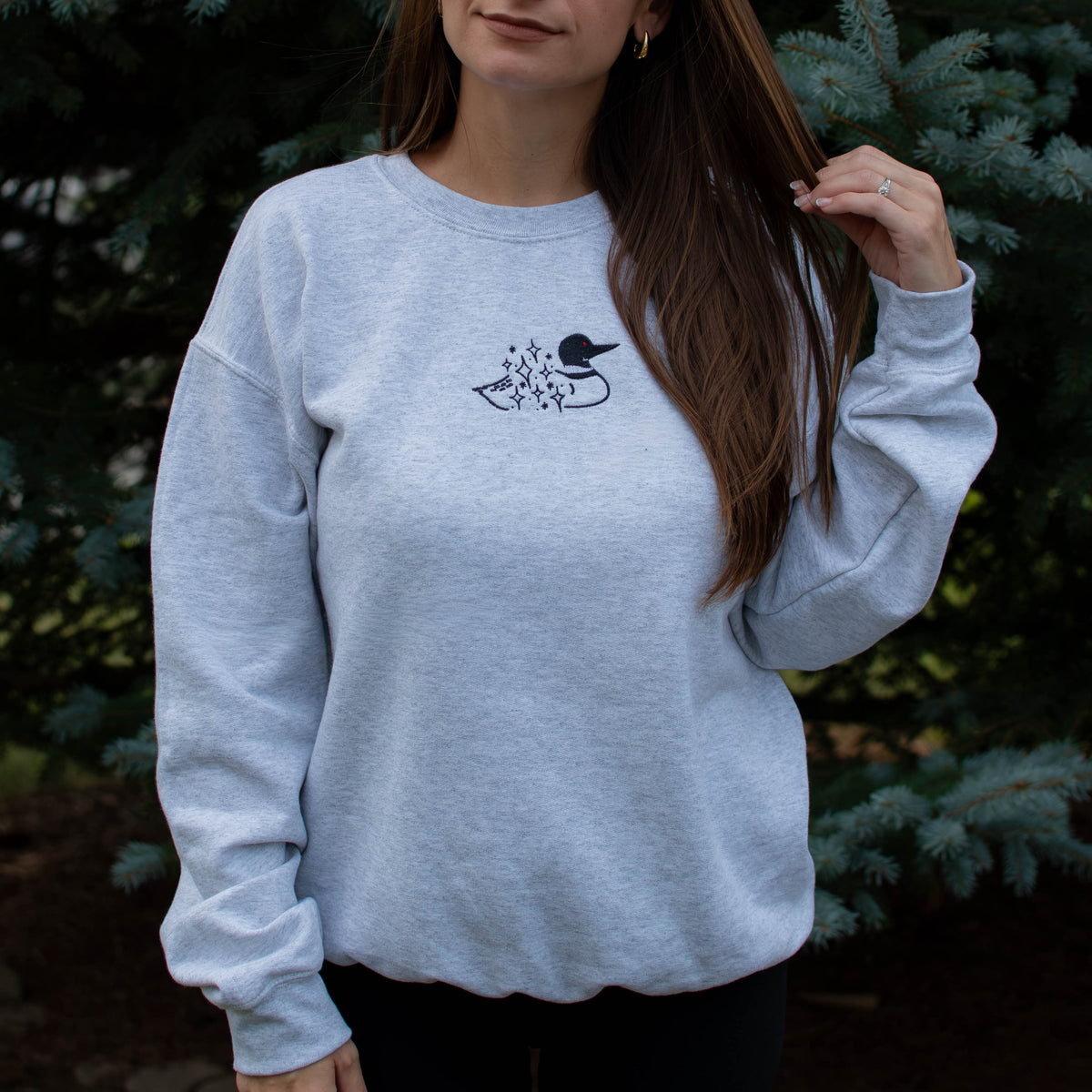 A woman wearing an ash colored crewneck sweatshirt with a small black embroidered loon design on the center chest, standing outdoors in front of green pine trees.
