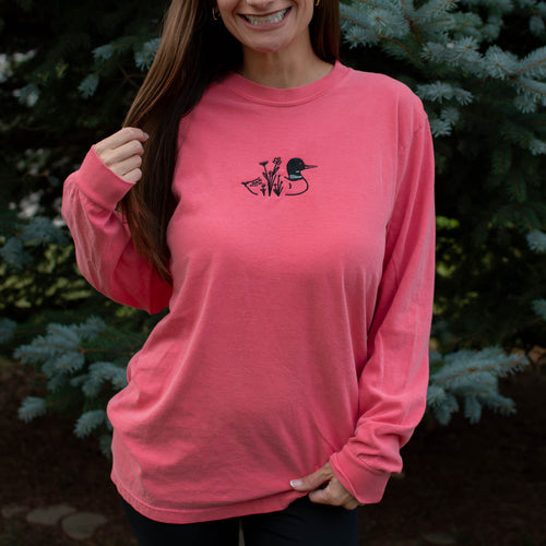 A woman wearing a watermelon colored crewneck sweatshirt with a small black embroidered loon design on the center chest, standing outdoors in front of green pine trees.