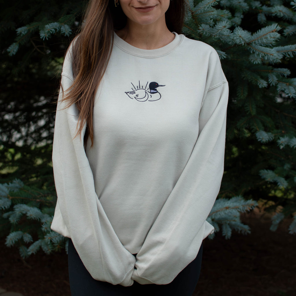 A woman wearing a sand colored crewneck sweatshirt with a small black embroidered loon design on the center chest, standing outdoors in front of green pine trees.