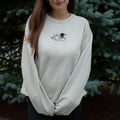 A woman wearing a sand colored crewneck sweatshirt with a small black embroidered loon design on the center chest, standing outdoors in front of green pine trees.