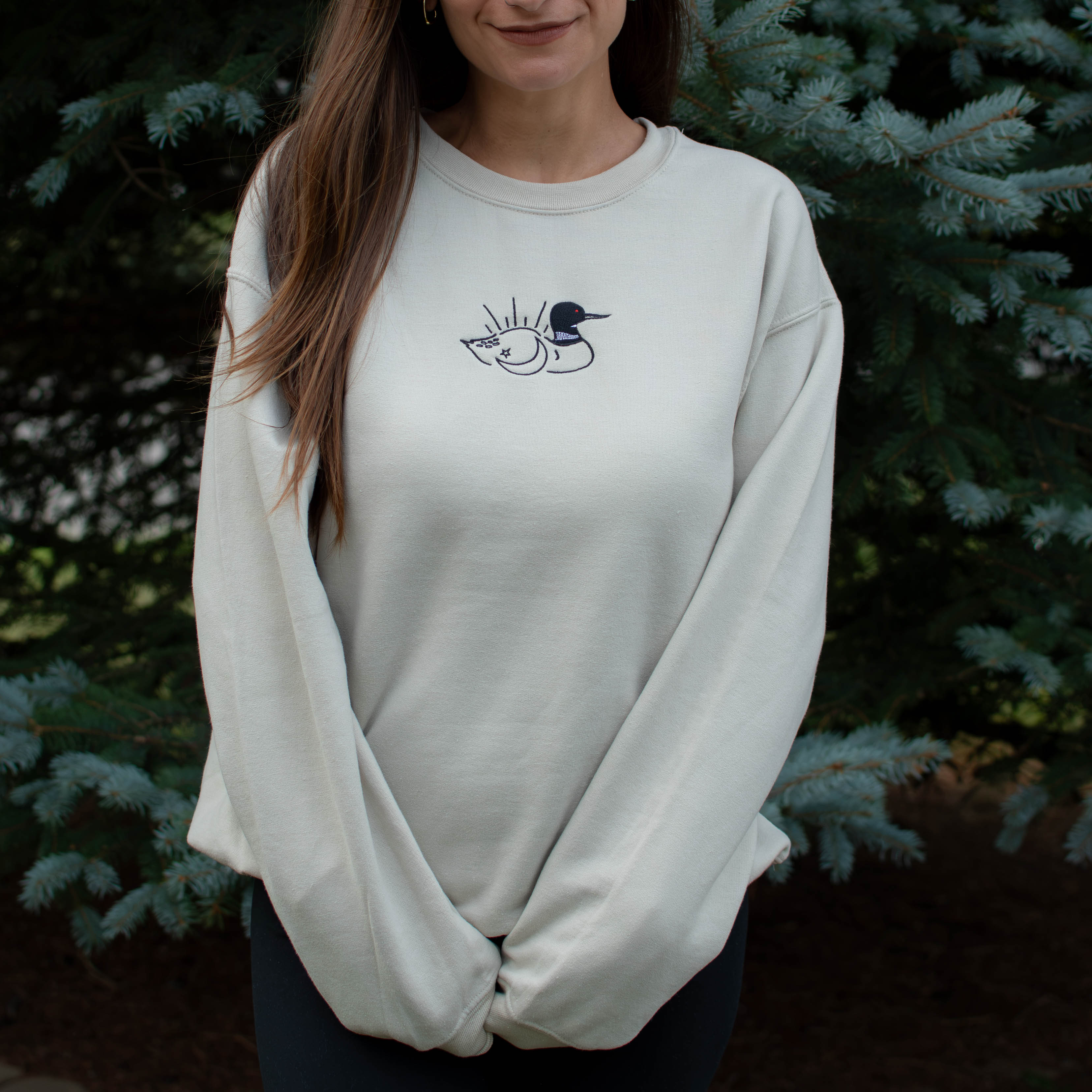 A woman wearing a sand colored crewneck sweatshirt with a small black embroidered loon design on the center chest, standing outdoors in front of green pine trees.