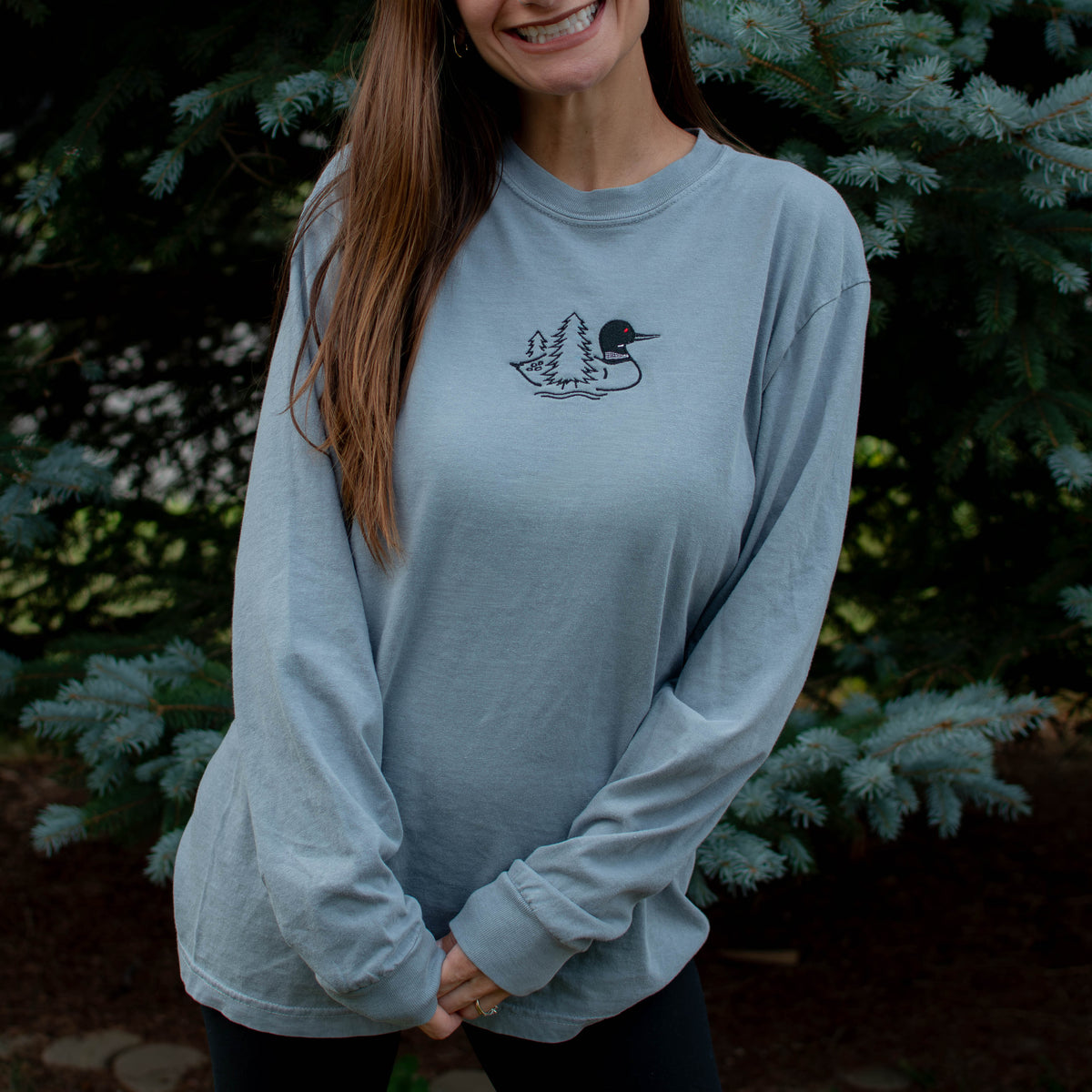 A woman wearing a granite colored crewneck sweatshirt with a small black embroidered loon design on the center chest, standing outdoors in front of green pine trees.
