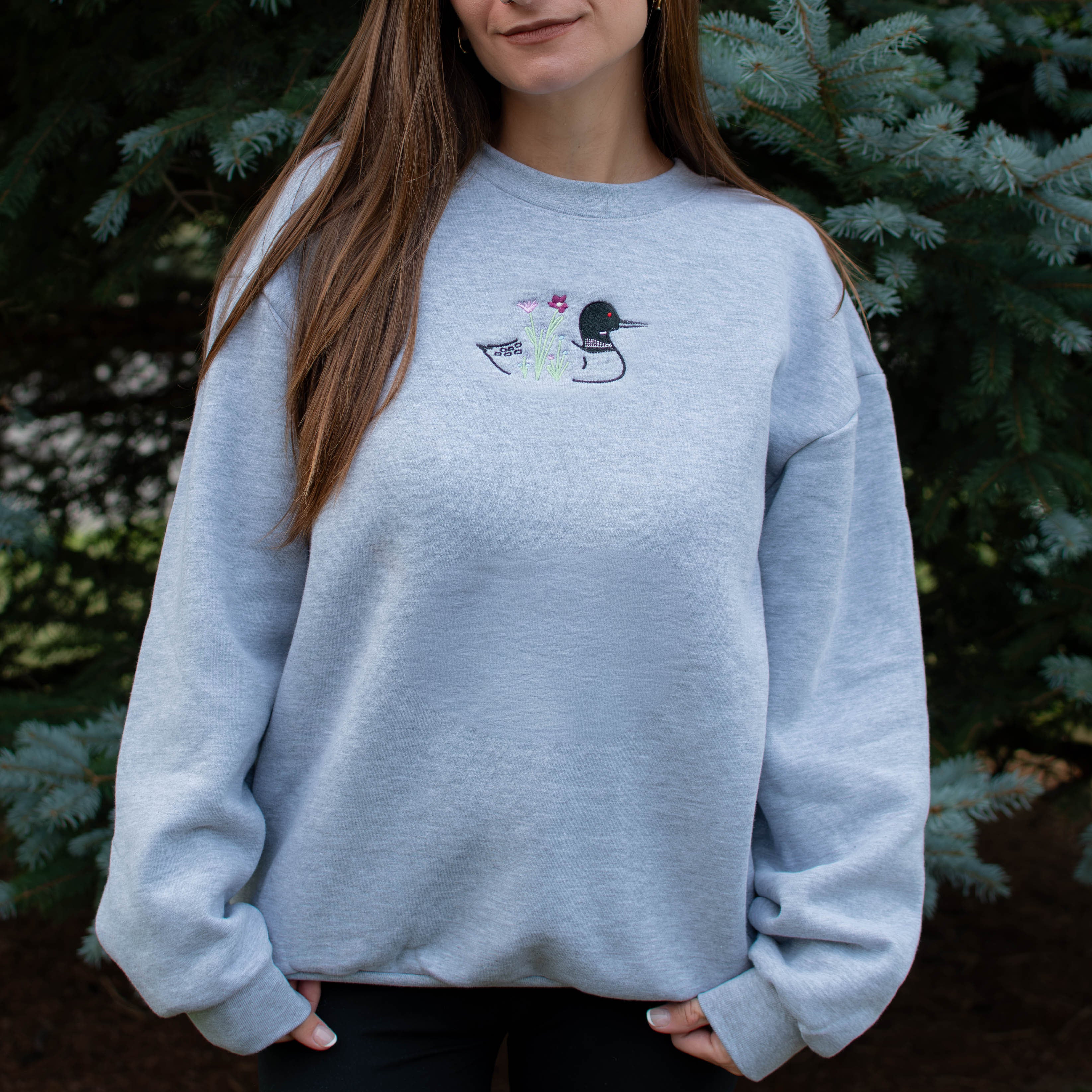 A woman wearing a light heather gray crewneck sweatshirt with a small embroidered loon design on the center chest, standing outdoors in front of green pine trees.