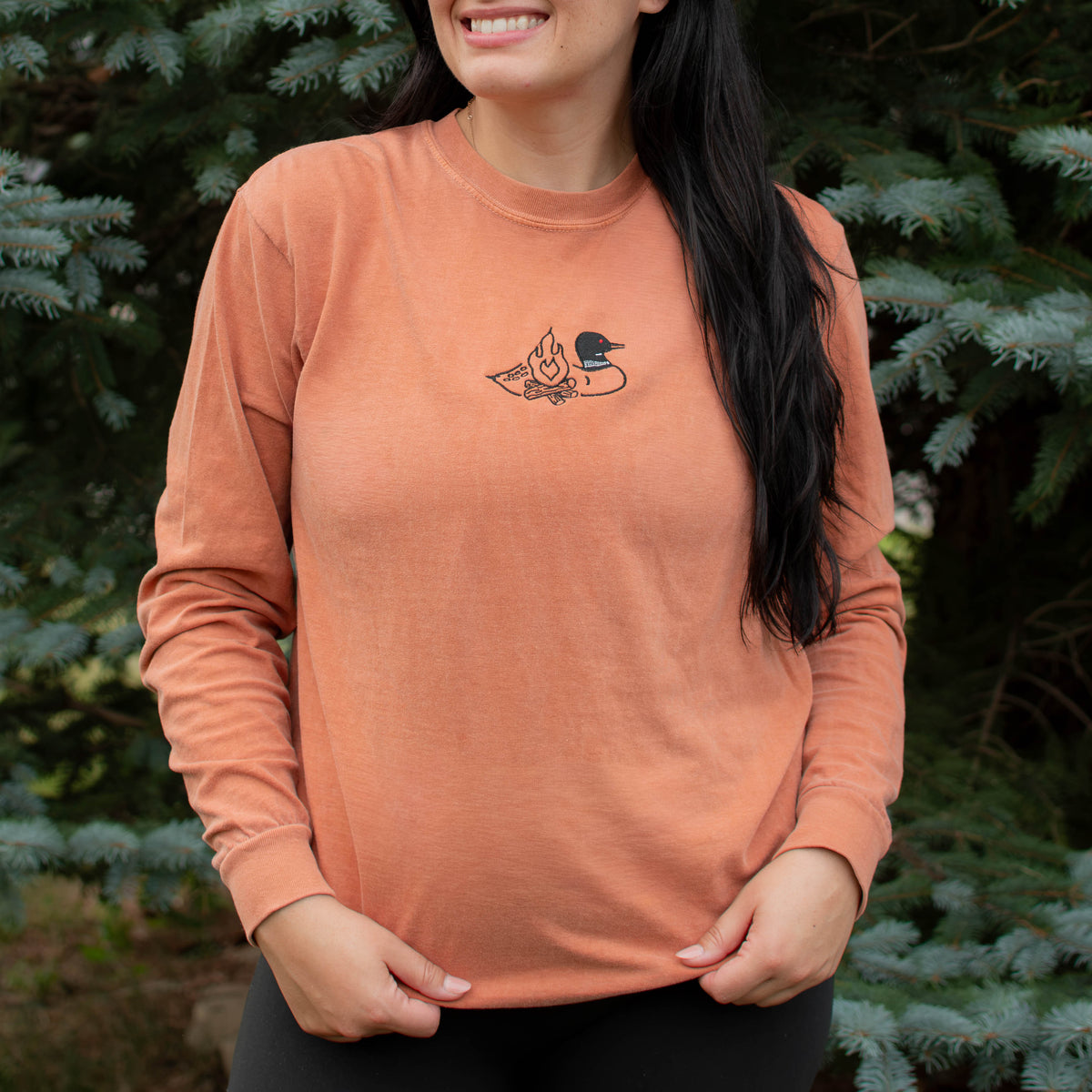 A woman wearing a yam colored crewneck sweatshirt with a small black embroidered loon design on the center chest, standing outdoors in front of green pine trees.
