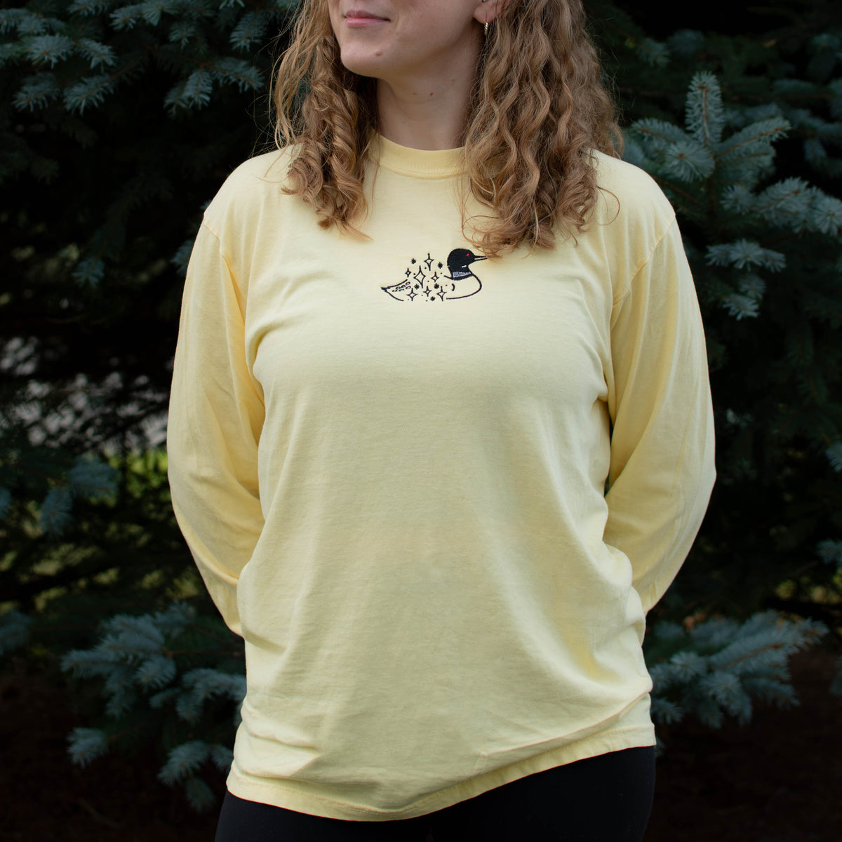 A woman wearing a butter colored crewneck sweatshirt with a small black embroidered loon design on the center chest, standing outdoors in front of green pine trees.
