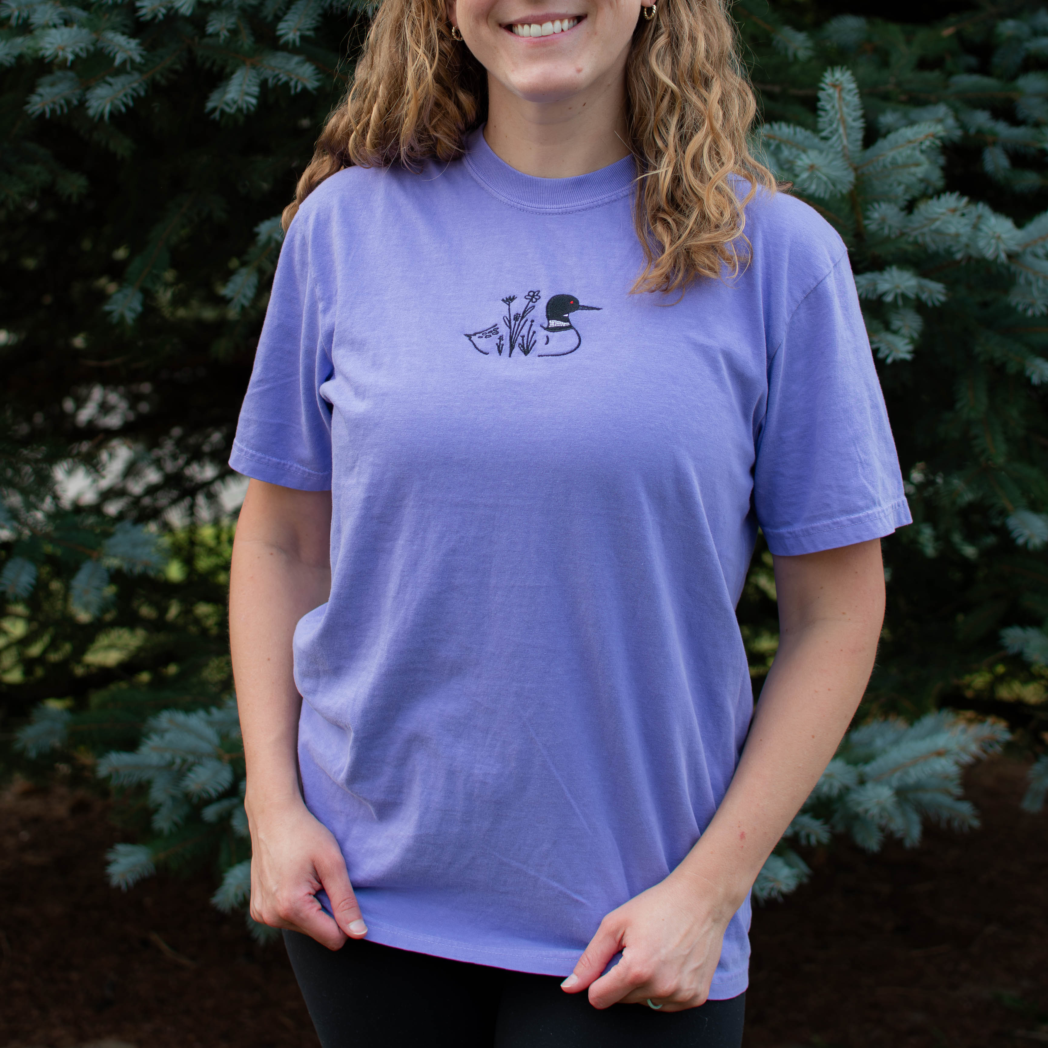A woman wearing a violet colored crewneck sweatshirt with a small black embroidered loon design on the center chest, standing outdoors in front of green pine trees.