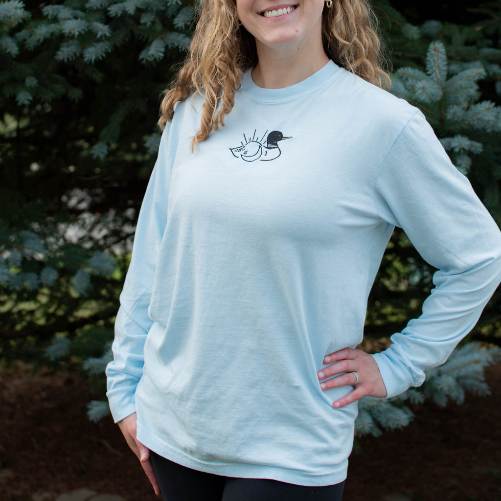 A woman wearing a chambray colored crewneck sweatshirt with a small black embroidered loon design on the center chest, standing outdoors in front of green pine trees.