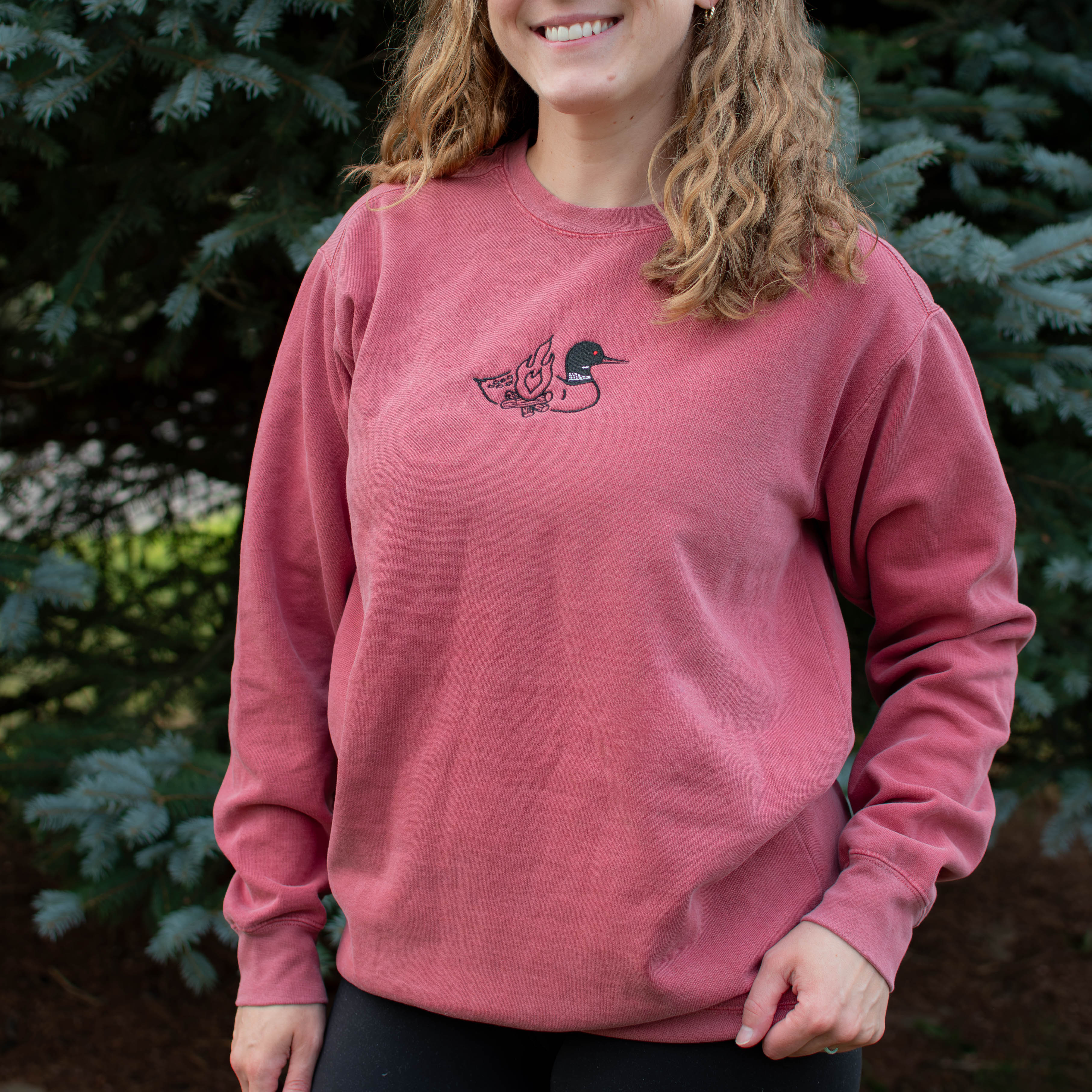 A woman wearing a crimson colored crewneck sweatshirt with a small black embroidered loon design on the center chest, standing outdoors in front of green pine trees.