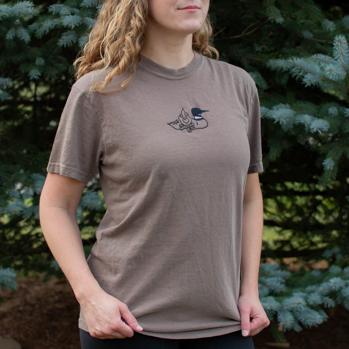 A woman wearing an espresso colored crewneck sweatshirt with a small black embroidered loon design on the center chest, standing outdoors in front of green pine trees.