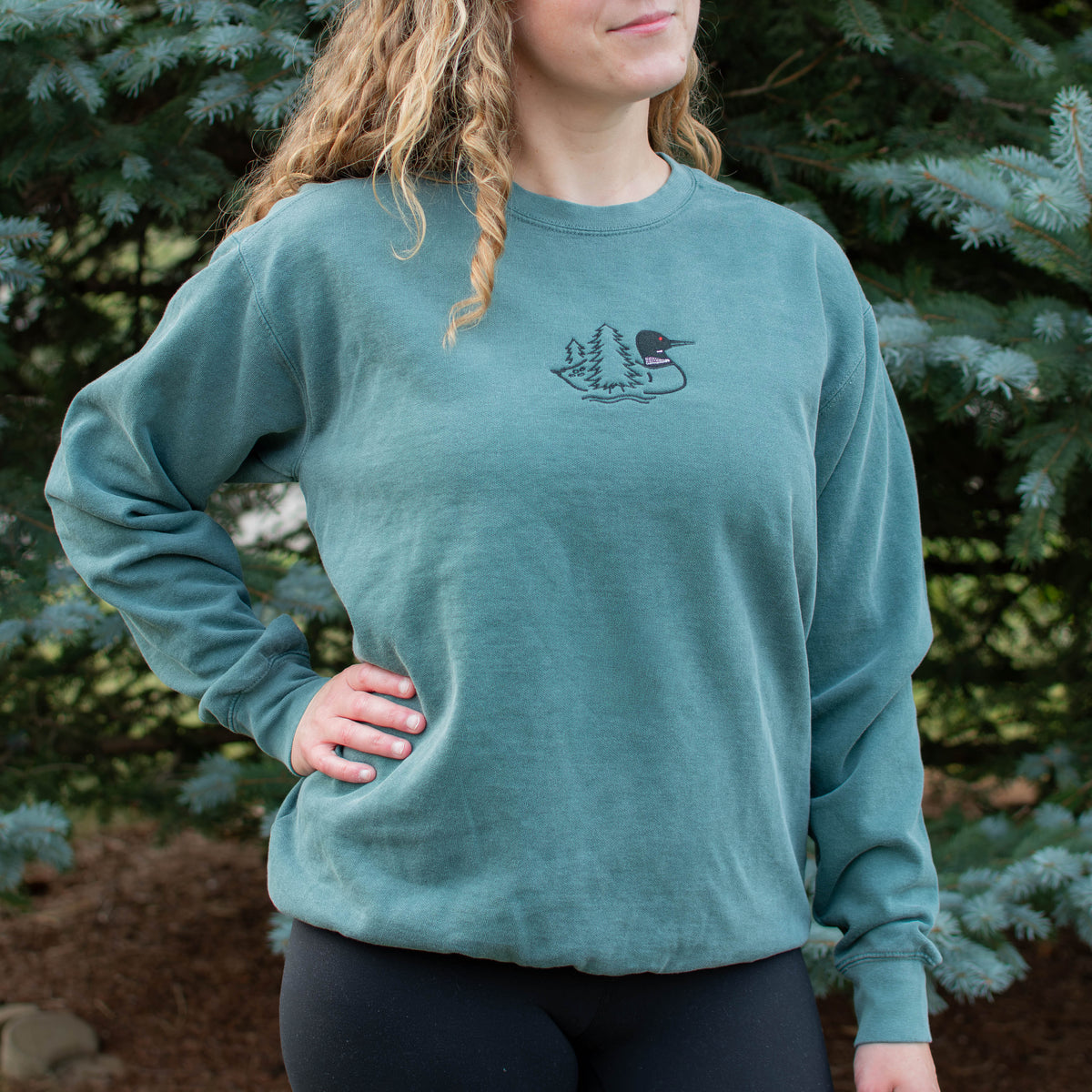 A woman wearing a blue spruce colored crewneck sweatshirt with a small black embroidered loon design on the center chest, standing outdoors in front of green pine trees.