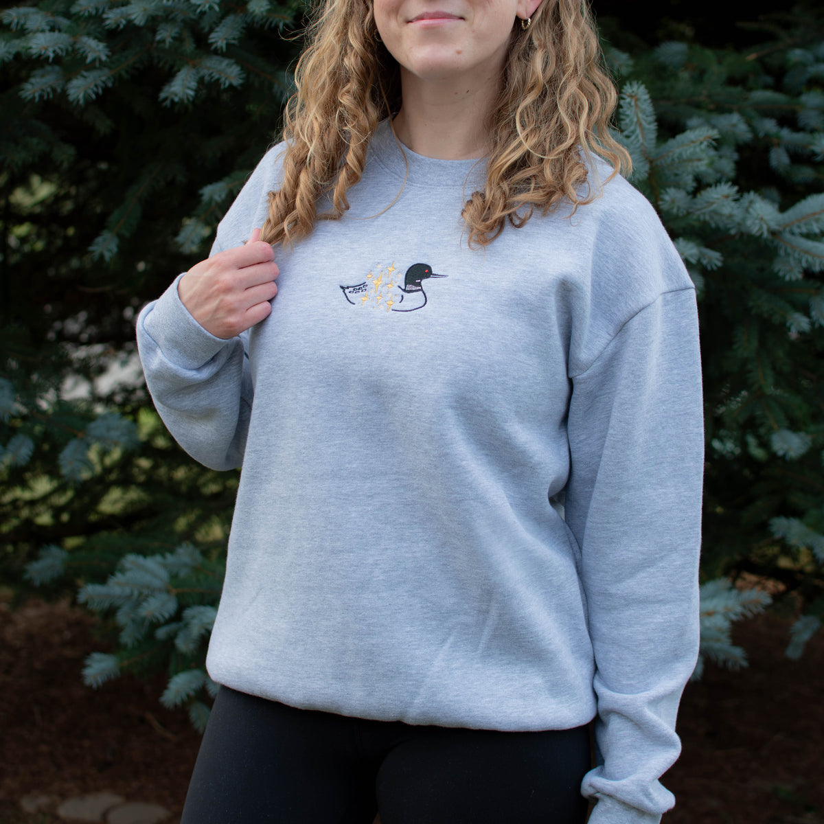 A woman wearing a light heather gray crewneck sweatshirt with a small embroidered loon design on the center chest, standing outdoors in front of green pine trees.
