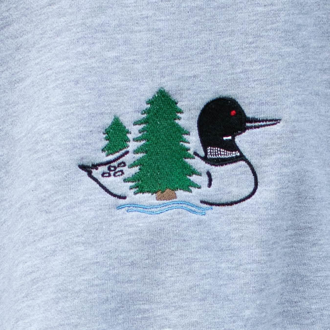 Close-up of a light heather gray fabric featuring an embroidered loon design with two green evergreen trees and wavy blue water with a small red eye detail.