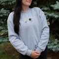 A woman wearing a light heather gray crewneck sweatshirt with a small embroidered loon design on the center chest, standing outdoors in front of green pine trees.