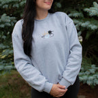 A woman wearing a light heather gray crewneck sweatshirt with a small embroidered loon design on the center chest, standing outdoors in front of green pine trees.
