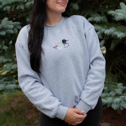 A woman wearing a light heather gray crewneck sweatshirt with a small embroidered loon design on the center chest, standing outdoors in front of green pine trees.