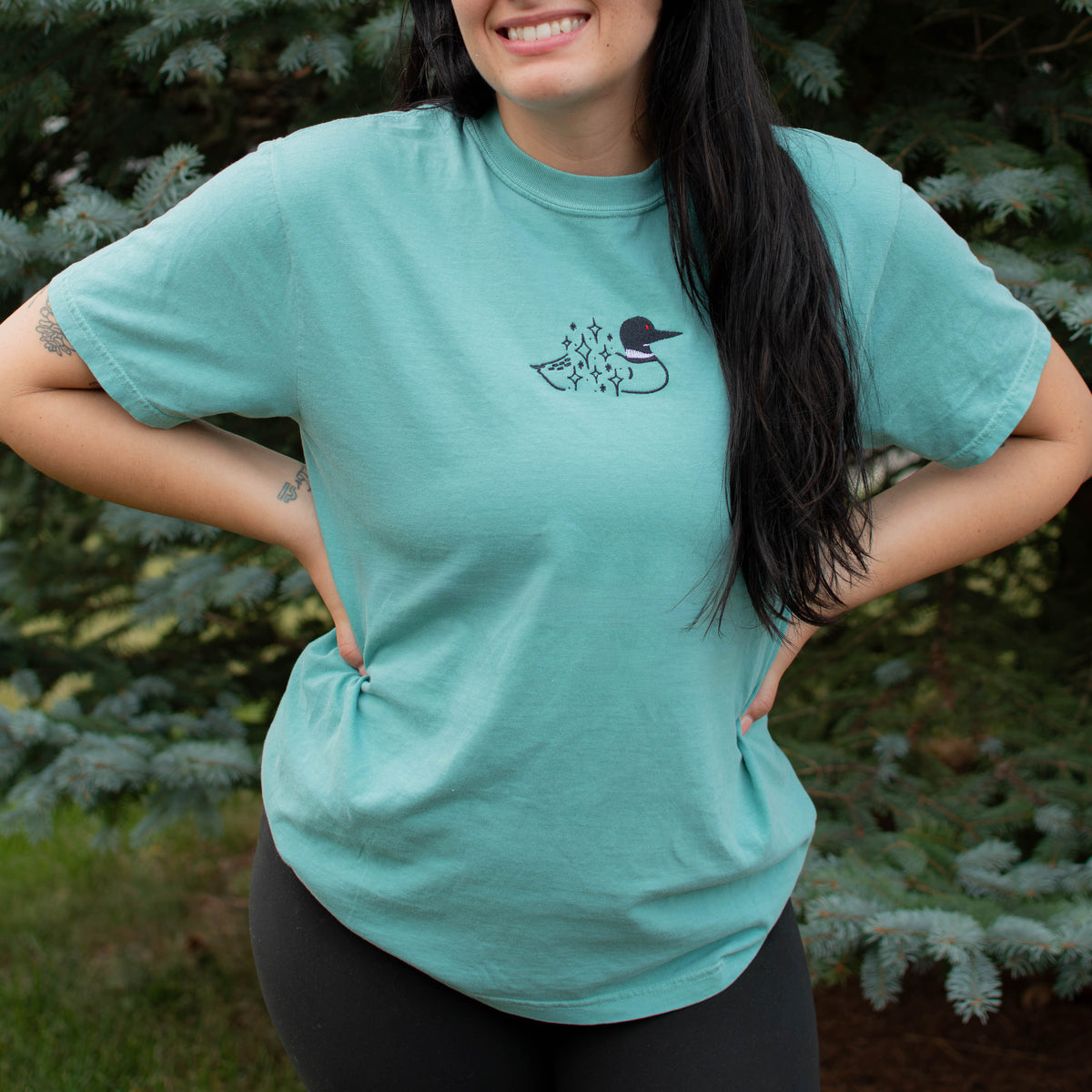 A woman wearing a seafoam colored crewneck sweatshirt with a small black embroidered loon design on the center chest, standing outdoors in front of green pine trees.