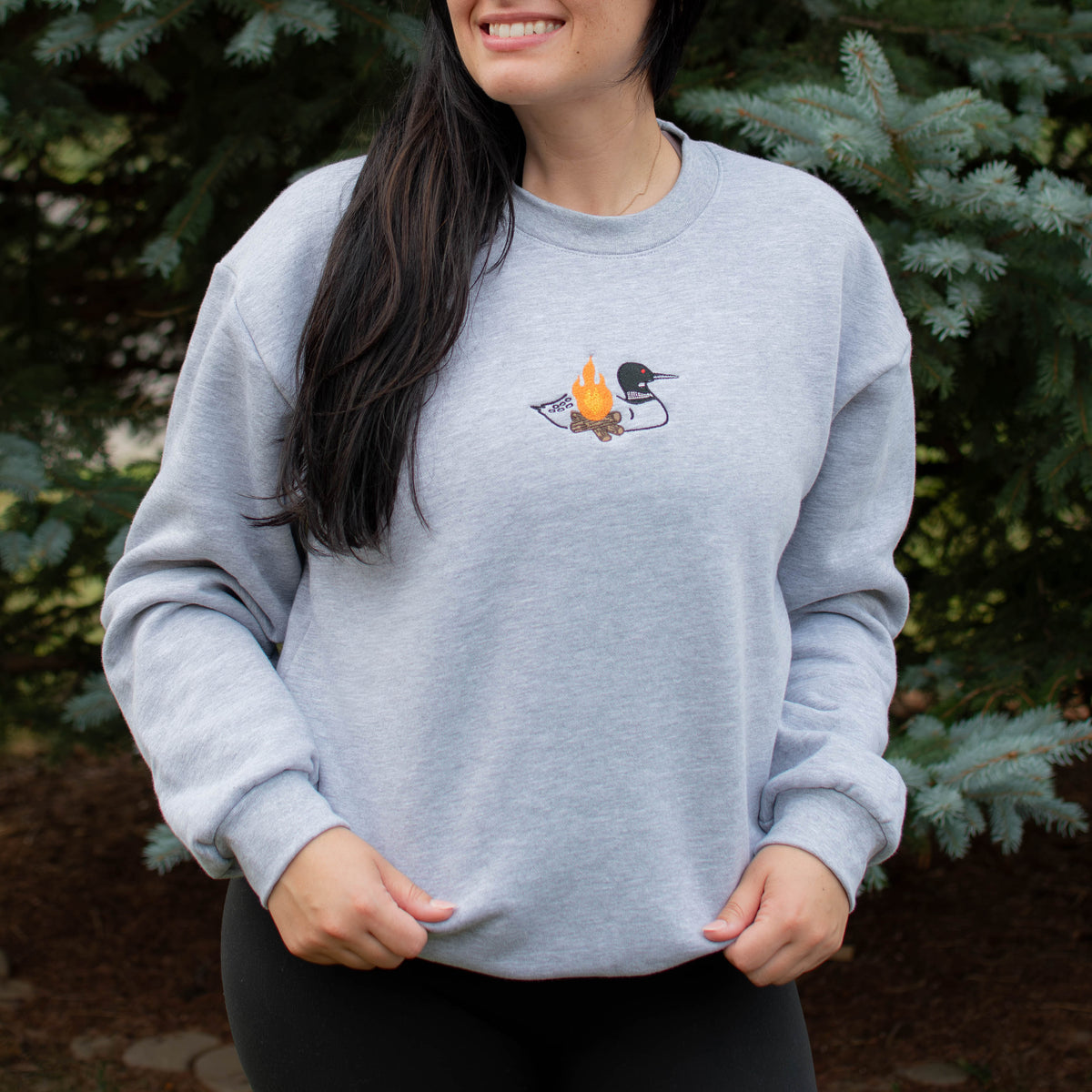A woman wearing a light heather gray crewneck sweatshirt with a small embroidered loon design on the center chest, standing outdoors in front of green pine trees.