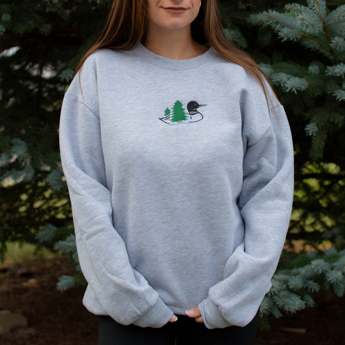 A woman wearing a light heather gray crewneck sweatshirt with a small embroidered loon design on the center chest, standing outdoors in front of green pine trees.