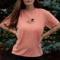 A woman wearing a terracotta colored crewneck sweatshirt with a small black embroidered loon design on the center chest, standing outdoors in front of green pine trees.