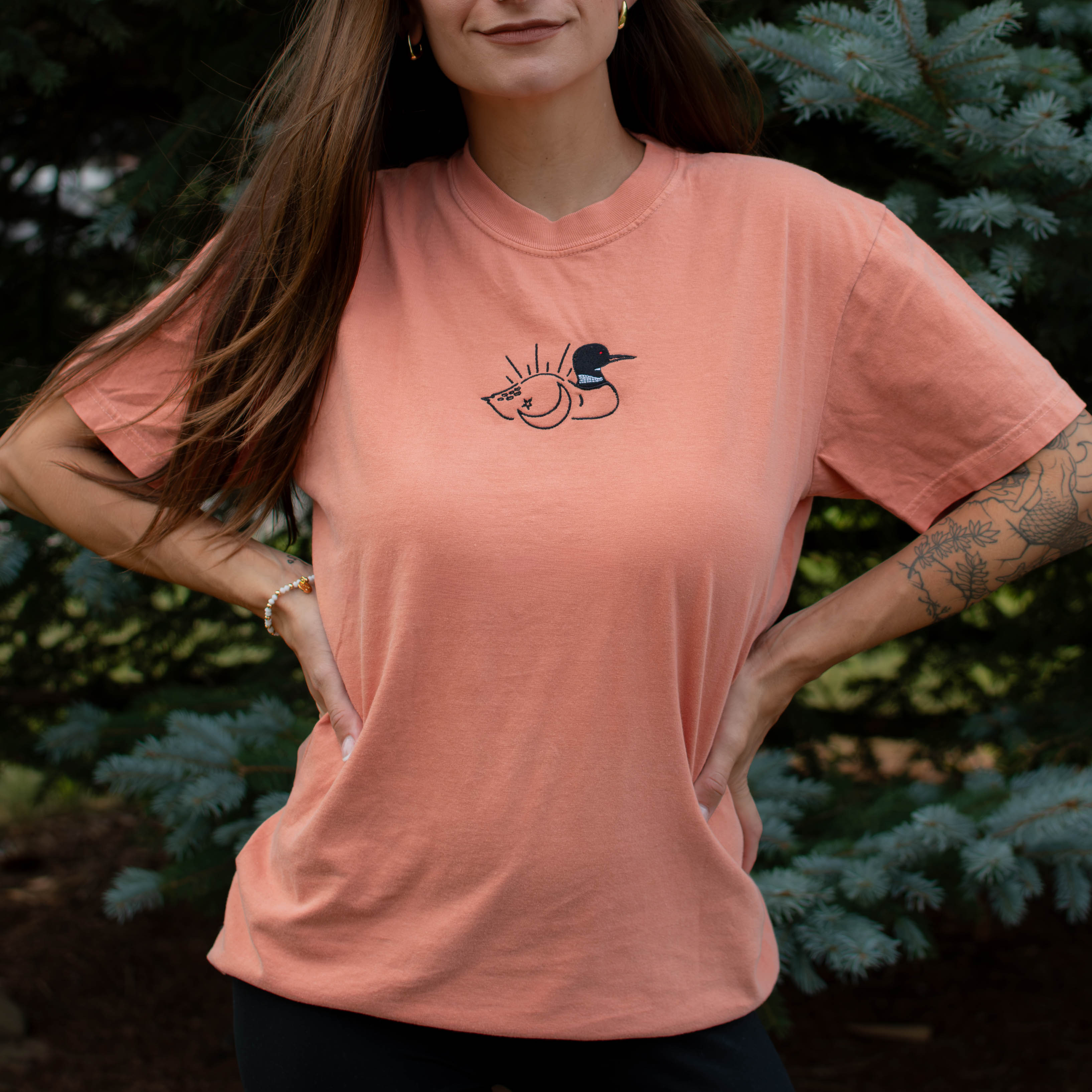 A woman wearing a terracotta colored crewneck sweatshirt with a small black embroidered loon design on the center chest, standing outdoors in front of green pine trees.